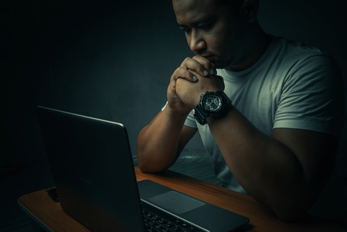A man sitting in a dark room with his hands clasped in front of a laptop, symbolizing investigations and allegations handled by a Tampa internet sex crimes lawyer.