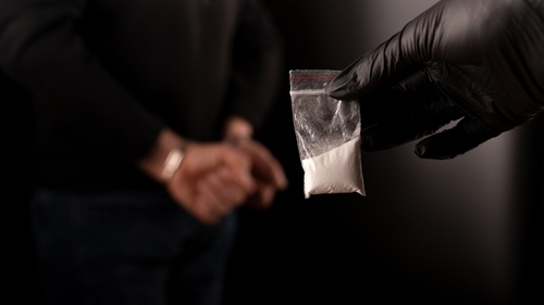 Gloved hand holding a small bag of white powder while a handcuffed person stands in the background, illustrating cases involving substances classified under the Florida Drug Schedule.